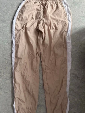 Beige mjukisbyxor med vita ränder - Snygga beige byxor från Better Together i 100% nylon. De har vita ränder längs sidorna och resår i midjan för en skön och avslappnad passform. Perfekta för en chill och sportig look.