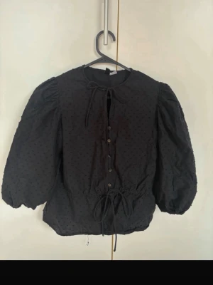 Svart blus med puffärm och knyt - Trendig svart blus med puffiga trekvartsärmar, knappar framtill och knytband i midjan och vid halsen. Tunt tyg med diskret prickstruktur ger en cool look. Perfekt för dig som gillar volym och detaljer.