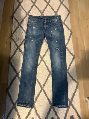 Dondup jeans med slitningar - Säljer ett par blå dondup jeans med slitningar och raka ben. Jeansen har klassisk femficksmodell och detaljerade slitningar framtill och baktill för en trendig look. Tillverkade i denim med en snygg tvättad finish. Slim/skinny fit