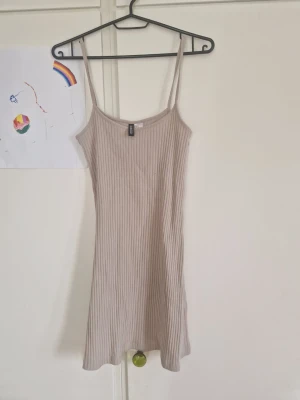 Beige ribbad kort klänning H&M - Säljer en beige ribbad kort klänning från H&M med smala axelband och figurnära passform. Perfekt för varma dagar och enkel att styla med olika accessoarer. Materialet är mjukt och stretchigt för extra komfort.