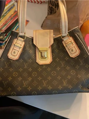 Monogram handväska - handväska i brunt monogramcanvas med beige läderdetaljer och guldiga metalldelar. Väskan har två rejäla handtag och ett lock med spänne framtill. Ikoniskt mönster över hela väskan och snygga kontrastsömmar. Lite ”sprucken” i materialet på beiga knapptygey se bild nummer 2 