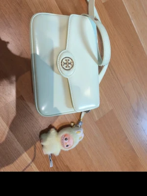 Gul axelremsväska från Tory Burch - den lilla Labubu ska följa med på köpet. Väskan har gott om plats för både en stor mobil och en liten sminknecessär.