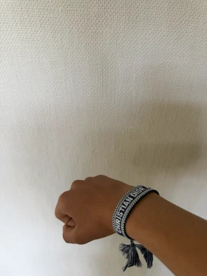 Dior armband - Säljer ett snyggt blått armband från Christian Dior med broderad logotyp och vita detaljer. Armbandet har justerbar knytning med tofsar och är gjort i textilmaterial. Perfekt för dig som vill ha en trendig accessoar med lyxig känsla.