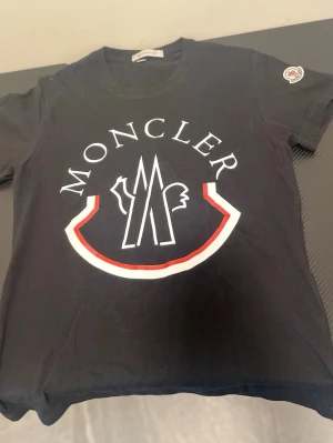 Svart Moncler t-shirt med stor logga - Snygg svart t-shirt från Moncler med stor vit och röd logga framtill. Klassisk rund halsringning och korta ärmar. Tillverkad i mjuk bomull och har en Moncler-patch på ena ärmen. Perfekt för dig som gillar streetwear och exklusiva märken.