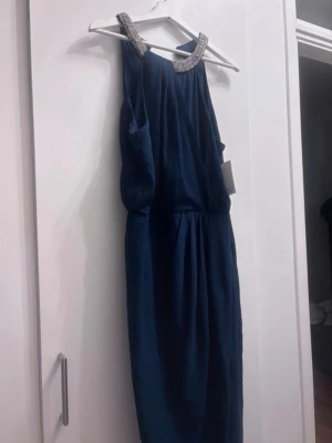 Marinblå långklänning med pärldetaljer - Elegant marinblå långklänning med pärlbroderad halsringning. Klänningen har ett löst fall och är ärmlös, vilket ger en stilren och festlig look. Perfekt för speciella tillfällen där du vill sticka ut med snygga detaljer. Xxl