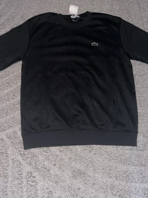 Svart stickad tröja från Lacoste - Klassisk svart stickad tröja från Lacoste med rund halsringning och den ikoniska gröna krokodilloggan broderad på bröstet. Tröjan har långa ärmar och ribbade muddar vid ärmslut och nederkant. Perfekt för en stilren och enkel look.