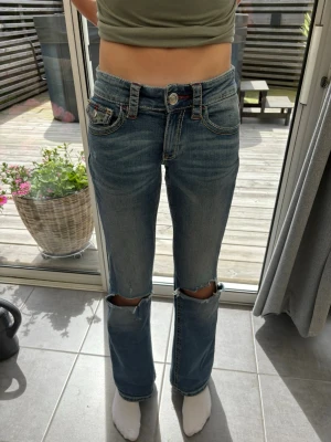 Blå lågmidjade jeans med hål på knäna - Säljer ett par blå jeans med hål på knäna och snygga detaljer på bakfickorna. Jeansen har låg midja, raka ben och kontrastsömmar. Materialet är klassisk denim och de har coola fickor både fram och bak med röda broderier.