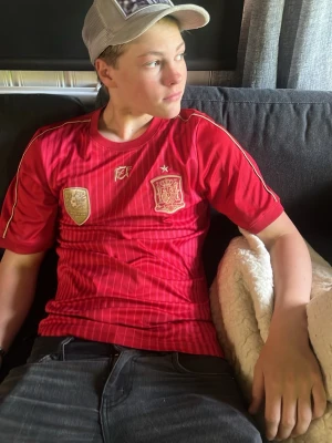 Röd Spanien fotbollströja Adidas - Säljer en röd Spanien fotbollströja från Adidas med tunna vita ränder, guldiga detaljer och klubbmärke på bröstet. Tröjan har korta ärmar och rund hals. Perfekt för dig som älskar fotboll och vill stötta landslaget.