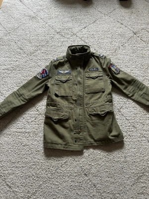 Olivgrön vindjacka Polo Ralph Lauren - Snygg olivgrön vindjacka från Polo Ralph Lauren Denim & Supply med coola patchar på ärmarna och bröstet. Jackan har fyra fickor framtill, hög krage och detaljerade broderier. Tillverkad i bomull för en skön och avslappnad look. Storlek 10-12 