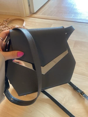 RULA GALAYANI - Svart geometrisk axelväska i skinn - Snygg svart axelväska i ett geometriskt snitt med silverdetaljer och justerbar axelrem. Väskan är gjord i strukturerat skinn och har en unik design med metallisk V-formad detalj på framsidan samt nitar på baksidan. Perfekt för dig som gillar statement-accessoarer.