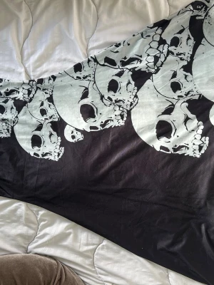 Svart långklänning med dödskallar - Unik svart långklänning med stora vita dödskalletryck längs nederkanten. Klänningen har en alternativ och edgy stil, perfekt för dig som vill sticka ut. Materialet känns mjukt och stretchigt, troligen bomull eller bomullsblandning.