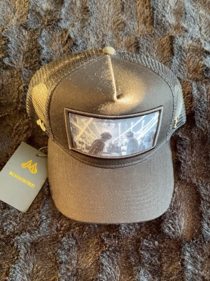 Svart truckerkeps från Maggiore - Snygg svart truckerkeps från Maggiore med mesh på sidorna och bak. Framtill finns en stor patch med svartvit foto-tryck av två personer i en urban miljö. Justerbar snapback och böjd skärm. Perfekt för dig som vill ha en unik och trendig accessoar.