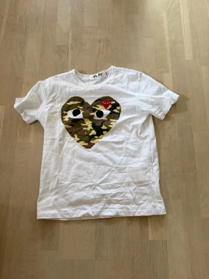 Vit Comme des Garçons PLAY t-shirt - Vit t-shirt från Comme des Garçons PLAY med stort hjärta i camouflage-mönster och ett litet rött hjärta med ögon på bröstet. Klassisk rund hals och korta ärmar. Perfekt statement-plagg för dig som gillar streetwear.