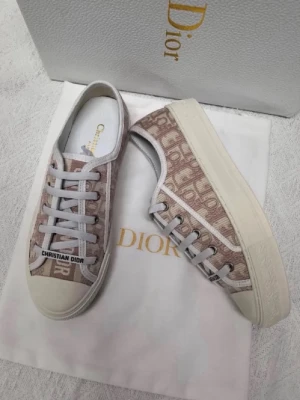 Beige Dior sneakers med monogram - Snygga sneakers från Christian Dior i beige och vitt med ikoniskt Dior-monogram på sidan. Skorna har vita snören, gummisula och detaljer i canvas. Perfekta för dig som vill ha en exklusiv touch till din outfit.
