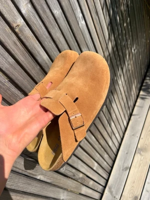 Birkenstocks - Säljer ett par snygga bruna tofflor från Birkenstock i mjuk mocka. Modellen har en bred rem med metallspänne och öppen häl, perfekt för chill dagar. Klassisk korksula som formar sig efter foten och ger grym komfort. Ser typ nya ut och de är soft footbed modellen så dem är väldigt mjuka och sköna