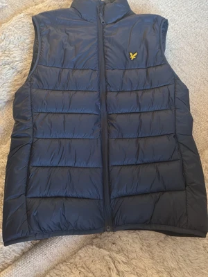 Mörkblå dunväst från Lyle & Scott - Snygg mörkblå dunväst från Lyle & Scott med gul logotyp på bröstet. Västen har ståkrage, dragkedja framtill och är quiltad med horisontella sömmar. Perfekt för lager-på-lager och håller dig varm under kyliga dagar.