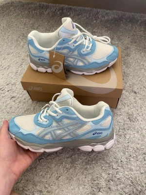 Asics vita och ljusblå sneakers - Snygga sneakers från Asics i vitt och ljusblått med mesh och syntetmaterial. Skorna har en chunky sula och klassisk Asics-logga på sidan. Perfekta för dig som gillar sportig stil och vill ha bekväma skor till vardags.
