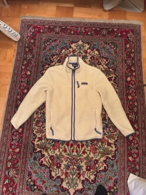 Beige teddyjacka från Patagonia - Säljer en beige teddyjacka från Patagonia med blå detaljer. Jackan har hög krage, dragkedja framtill och två fickor med dragkedja. Mysigt och fluffigt material som håller dig varm. Klassisk och stilren design med logga på bröstet. Nypris: 1500kr
