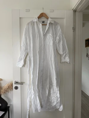 Vit randig skjortklänning Gina Tricot - Vit lång skjortklänning från Gina Tricot med tunna svarta ränder. Klänningen har lång ärm, krage, knappar framtill och ett bälte i midjan. Perfekt för en avslappnad och stilren look. Vid fästet för bälten har ena sidan lossnat av, upptäckte det efter att jag har köpt den. Låg i garderoben och hade helt glömt bort om den, därför väljer jag att sälja den för ett billigare pris än vad jag fick köpa den för! 
