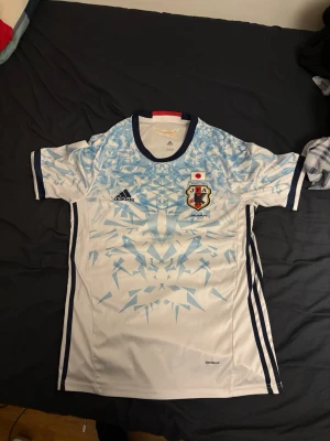 Japans landslagströja Adidas - Säljer en vit och ljusblå Japans landslagströja från Adidas med geometriskt mönster upptill, mörkblå detaljer och rund hals. Tröjan har korta ärmar och är tillverkad i lätt och ventilerande polyester, perfekt för fotboll eller träning. De finns små fläckar med svart där bak syns knappast 