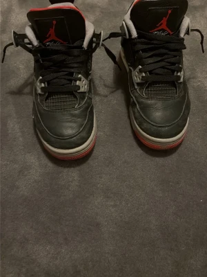 Svarta Air Jordan 4 Retro sneakers - Säljer ett par klassiska Air Jordan 4 Retro sneakers i svart läder med grå och röda detaljer. Skorna har den ikoniska Jumpman-loggan på plösen och sulan, samt meshpaneler och svarta skosnören. Perfekta för dig som gillar streetwear och snygga detaljer.