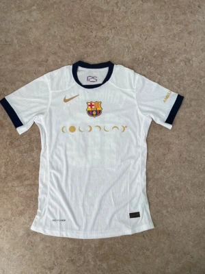 Vit FC Barcelona matchtröja Nike - Snygg vit FC Barcelona matchtröja med mörkblå detaljer och guldfärgade tryck. Tröjan har klubbmärke, Nike-logga och månfas-tryck framtill samt Lamine Yamal och nummer 10 på ryggen. Tillverkad i lätt och ventilerande material, perfekt för fotbollsträning.