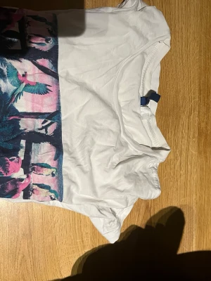 Vit t-shirt med färgglatt tryck - Vit t-shirt från Divided med ett stort färgglatt tryck framtill som visar papegojor och palmer i blått, rosa och grönt. Klassisk rund hals och korta ärmar. Tillverkad i mjuk bomull som känns skön mot huden. Perfekt för dig som gillar statement-plagg.