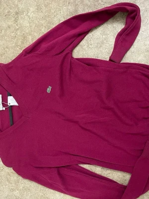 Vinröd stickad tröja från Lacoste - Vinröd stickad tröja från Lacoste med klassisk v-ringning och den ikoniska gröna krokodilloggan på bröstet. Tröjan har långa ärmar och ribbade muddar vid ärmslut och nederkant. Perfekt för dig som gillar stilrena och tidlösa plagg.
