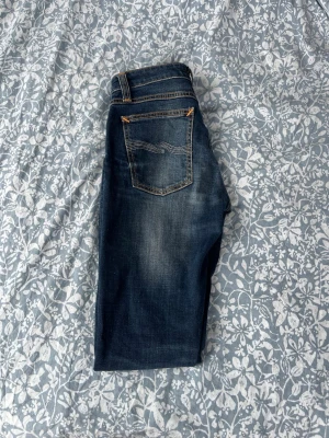Mörkblå skinny jeans från Nudie - Säljer ett par mörkblå skinny jeans från Nudie med snygga slitningar och kontrastsömmar. Klassisk femficksmodell med läderpatch bak i midjan och broderade bakfickor. Perfekta för dig som gillar en tight passform och streetig look.