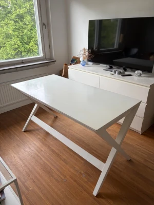 Stilrent vitt skrivbord med X-ben - Säljer ett modernt skrivbord i vitt med rektangulär bordsskiva och snygga X-formade ben. Bordet är tillverkat i trä eller träkomposit och har en minimalistisk design som passar perfekt till plugghörnan eller hemmakontoret.