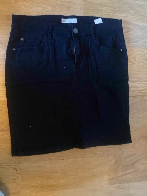 Svart jeanskjol med fickor - Snygg svart jeanskjol med klassisk femficksmodell och dragkedja framtill. Kjolen har bälteshällor och raka linjer för en stilren look. Perfekt att matcha med en t-shirt eller hoodie för en avslappnad vibe.