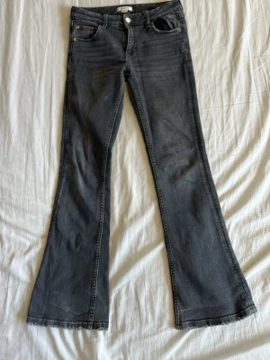 Grå bootcut jeans med rå kant - Säljer ett par grå bootcut jeans med rå kant längst ner. Jeansen har klassiska fem fickor och en knappstängning framtill. Materialet är denim och passformen är utsvängd från knäna och ner. Perfekt för dig som gillar en retrostil. De är ett litet märke i bak men de syns inte när man har på sig dom