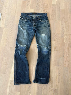 Galet snygga nudie jeans - Säljer ett par blå jeans med slitningar och hål på knäna samt slitna detaljer vid bensluten. Jeansen har fem fickor och klassisk passform. Perfekta för en avslappnad och trendig look. Fråga gärna om mått. 