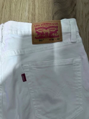  Levi's 511 - Säljer ett par vita Levi's 511 slim jeans med klassisk femficksdesign och röd Levi's-tag på bakfickan. Jeansen har normal midja och raka ben, tillverkade i bomull med en liten stretch för extra komfort.