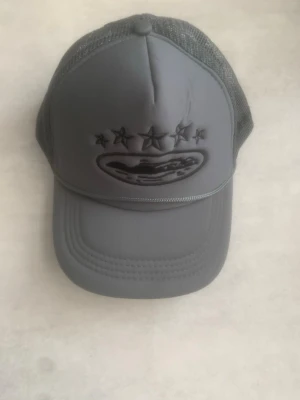 Grå keps - Snygg grå keps med mesh på sidorna och bak, samt broderade stjärnor och ett ovalt märke framtill. Kepsen har böjd skärm och justerbar baksida för perfekt passform. Perfekt för en streetwear-look.