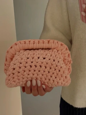 Handvirkad rosa clutchbag - Superfin handvirkad clutchbag i ljusrosa/ aprikos färg. Väskan har en mjuk, chunky struktur och är gjord av tjockt garn som ger en trendig och unik look. Perfekt accessoar för att lyfta din outfit med en söt och personlig touch.