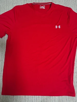 Röd t-shirt från Under Armour - Röd t-shirt från Under Armour med klassisk passform och korta ärmar. T-shirten har en liten vit logga på bröstet och är tillverkad i ett mjukt och lätt material som passar perfekt till träning eller vardag.