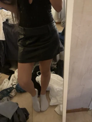 Svart kort skinnkjol - Säljer en svart kort kjol i skinnimitation med normal passform. Kjolen har bälteshällor i midjan och en stilren look som är lätt att matcha med olika toppar. Perfekt för dig som gillar en edgy och trendig stil.