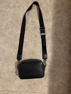 Svart axelväska från Valentino - Snygg svart axelväska från Valentino i skinn med justerbar rem där loggan är tryckt. Väskan har dragkedja upptill, extra fack med dragkedja på baksidan och stilren design. Perfekt för dig som vill ha en trendig och praktisk väska. Ny pris 1200 kr mitt pris 349, ända ”defekterna” är att den har rostat lite vid dragkedjorna. Kontakta mig för bilder på det!