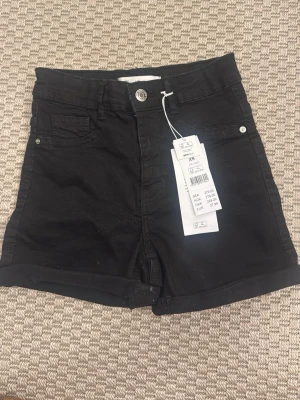 Svarta högmidjade shorts från Gina Tricot - Svarta högmidjade shorts från Gina Tricot med klassisk femficksdesign och uppvikta benslut. Stängs med knapp och dragkedja. Perfekta för en trendig och avslappnad look.