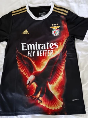 Benfica svart matchtröja Adidas - Snygg svart Benfica matchtröja från Adidas med guldiga axelränder och vit krage. Framsidan har ett stort tryck av en brinnande örn, klubbmärke och Emirates Fly Better-logga. Tillverkad i lätt och ventilerande material, perfekt för träning eller match.