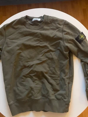 Olivgrön sweatshirt från Stone Island - Snygg olivgrön sweatshirt från Stone Island med klassisk rund hals och ikonisk patch på vänster ärm. Tröjan är tillverkad i mjuk bomull och har ribbade muddar vid ärmslut och nederkant. Perfekt för en avslappnad streetwear-look.