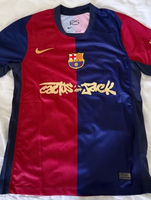 FC Barcelona x Cactus Jack matchtröja - Säljer en FC Barcelona matchtröja i samarbete med Cactus Jack och Nike. Tröjan är röd och marinblå med klubbmärke, Nike-logga och Cactus Jack-tryck framtill. Tillverkad i polyester med korta ärmar och sportig passform. Perfekt för fotboll eller som streetwear.