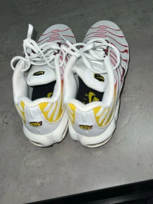 Nike Air Max Plus vita/röda/gula - Nike Air Max Plus sneakers i vitt med röda och gula detaljer. Skorna har det klassiska TN-märket på hälen, svarta swoosh-loggor på sidorna och synliga Air Max-enheter i sulan. Ovandelen är i mesh och syntet med vågformade linjer. Perfekta för dig som gillar en sportig och färgstark look.
