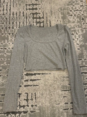 Grå croppad långärmad topp - Snygg grå croppad topp med lång ärm och rundad urringning. Toppen är ribbad och har en enkel, stilren design som passar perfekt till jeans eller kjol. Perfekt för dig som gillar en clean och trendig look.