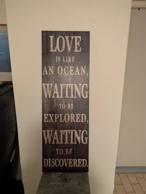 Dekorativ träskylt med citat - Snygg rektangulär träskylt i mörkbrunt med vitt tryck. Skylten har texten 'LOVE IS LIKE AN OCEAN, WAITING TO BE EXPLORED, WAITING TO BE DISCOVERED.' Perfekt som inredningsdetalj för att skapa en mysig och inspirerande känsla hemma.