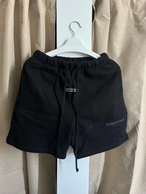 Essentials Fear Of God Shorts - Helt oanvända och fräscha sweat shorts från Essentials Fear of God i färgen svart. Endast uppackade för fotografering, taggen sitter fortfarande kvar. Perfekta för en snygg och bekväm streetwear-look. Egenskaper: • Märke: Essentials Fear of God (original). • Storlek: Small (S). Passformen är något oversized, som märket är känt för. • Färg: Svart. • Material: Kraftig och mjuk bomullsblandning (80% bomull, 20% polyester) med ruggad insida för maximal komfort.(Pris kan Diskuteras vid snabb affär!)
