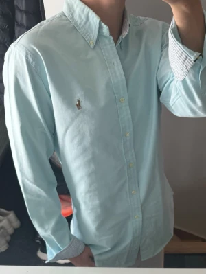 Ljusblå skjorta från Ralph Lauren - Klassisk ljusblå skjorta från Ralph Lauren med button-down krage och broderad logga på bröstet. Skjortan har långa ärmar och vita knappar framtill. Tillverkad i mjuk bomull och har en tidlös, clean look.