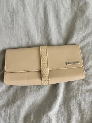 Beige plånbok från JANTAMINIAU - Stilren beige plånbok från JANTAMINIAU med magnetknäppning och strukturmönstrad rem. Plånboken har en metallbricka med logga på framsidan och KLM-tryck på insidan. 
