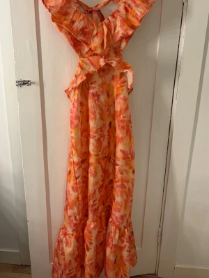 Färgglad långklänning med volanger - Säljer en långklänning i orange, rosa och gula toner med volangdetaljer och öppen rygg. Klänningen har ett unikt mönster och knytning i nacken, perfekt för sommarens alla tillfällen. Materialet känns lätt och luftigt.